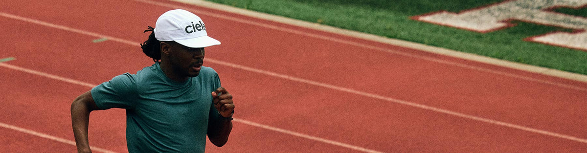 A man running on an all-weather track in a ciele athletics hat.