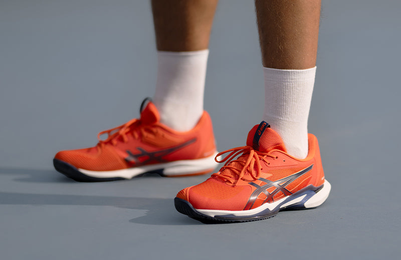 A man standing on a gray court in orange ASICS Solution Speed FF 3 tennis shoes.
