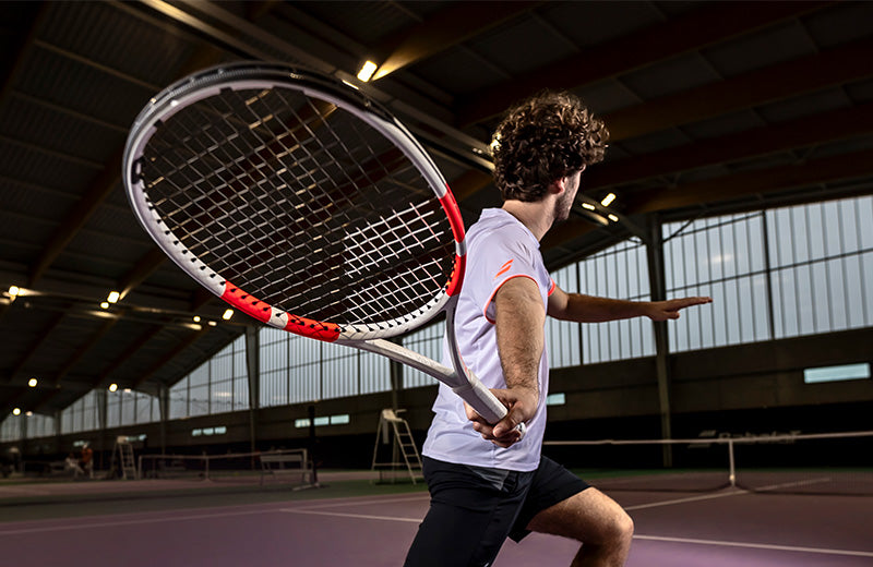 Lifestyle image: Mid-swing shot of a man playing with a Babolat Pure Strike 2024 tennis racquet on a dimly lit indoor court.