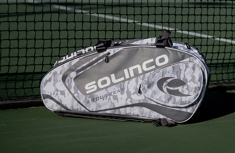 A white camo Solinco tennis bag sitting on a green tennis court.