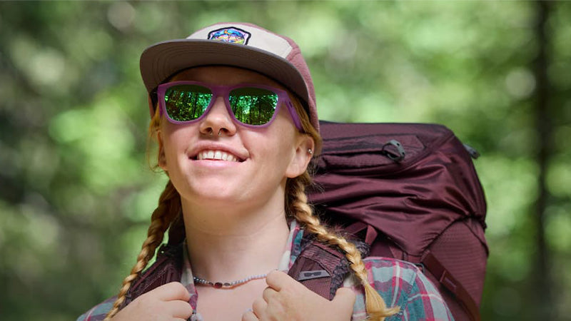 A person in a flannel shirt, trucker hat and purple goodr Sunglasses with green lenses wearing a large backpack in the woods with a blurred background
