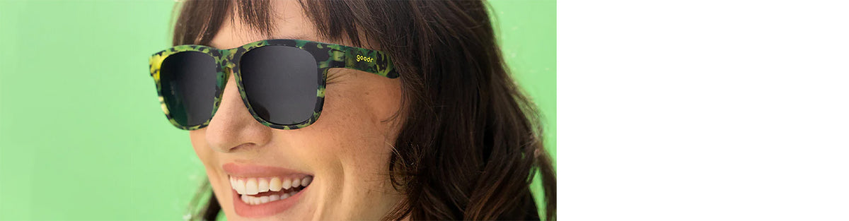 smiling woman weraing goodr howling at the neon moon bfg sunglasses on a green background