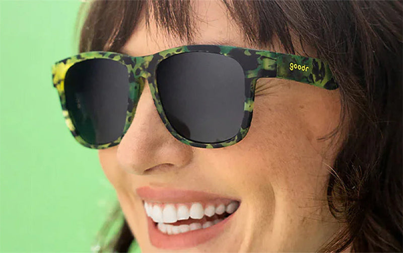 smiling woman weraing goodr howling at the neon moon bfg sunglasses on a green background