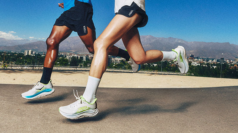 Two runners wearing HOKA Clifton 11 running shoes on a paved surface with a cityscape and mountains in the background.