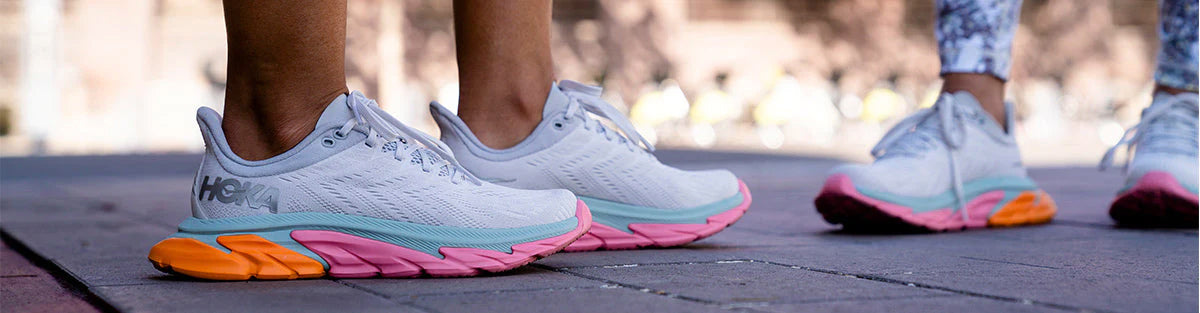 Two people standing on a paved area in light gray, light blue, neon orange and neon pink HOKA Clifton Edge running shoes.