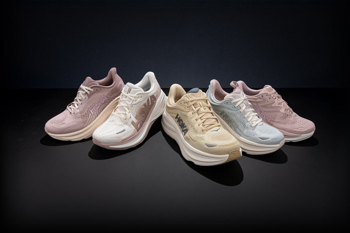 Five HOKA running shoes in soft pastel colors arranged on a smooth dark surface. The shoes include dusty pink, off-white, beige, and pale blue colors, with the HOKA logos clearly visible on the sides.