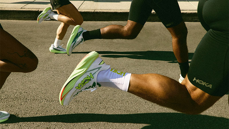 People running on a paved road in white HOKA Mach 7 running shoes with green and red accents