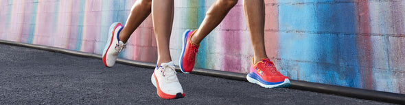 Man and woman in front of rainbow colored wall wearing HOKA Mach 4 Start Pack running shoes