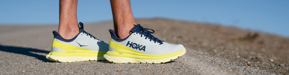 Man standing along a rural road wearing Hoka One One Mach 4 Blue Flower/Citrus running shoes.