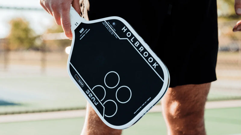 Closeup of a person holding a Holbrook Fuze pickleball paddle on a sunny court with a blury, washed out background