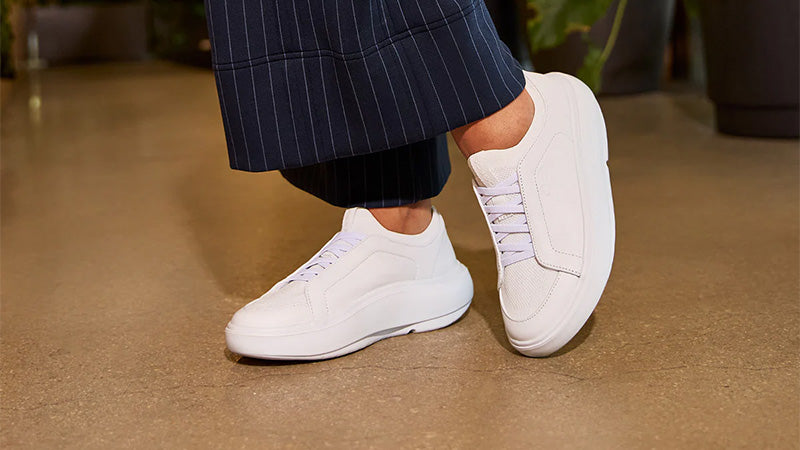 the feet of a person in dark blue pinstripe pants standing on a sand colored floor in white OOFOS Club+ walking shoes