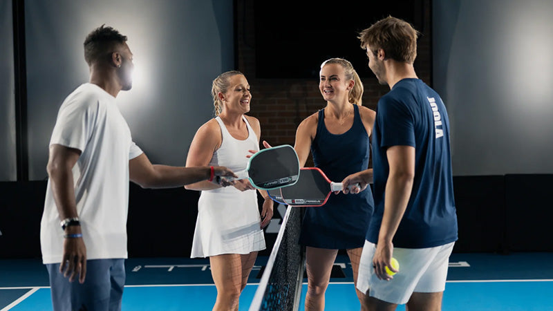 Four pickleball players meeting at the net on a blue court with JOOLA Pro V pickleball paddles in a mixed doubles match-up.