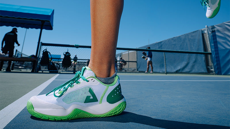 A pickleball player in motion wearing white and green JOOLA R4LLy Pickleball shoes on a blue court with other players and a clear blue sky in the background
