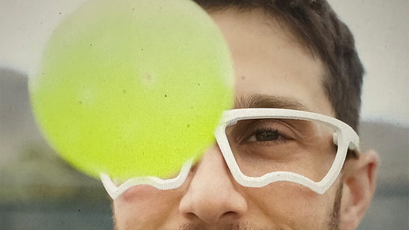 Closeup of a person wearing white Kitchen Blacker pickleball eyeguards with a blurred yellow pickleball in the foreground 