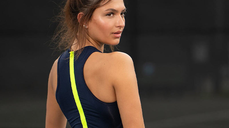 Woman wearing a dark blue Lucky in Love tennis dress with a bright yellow-green stripe down the back against a blurred background