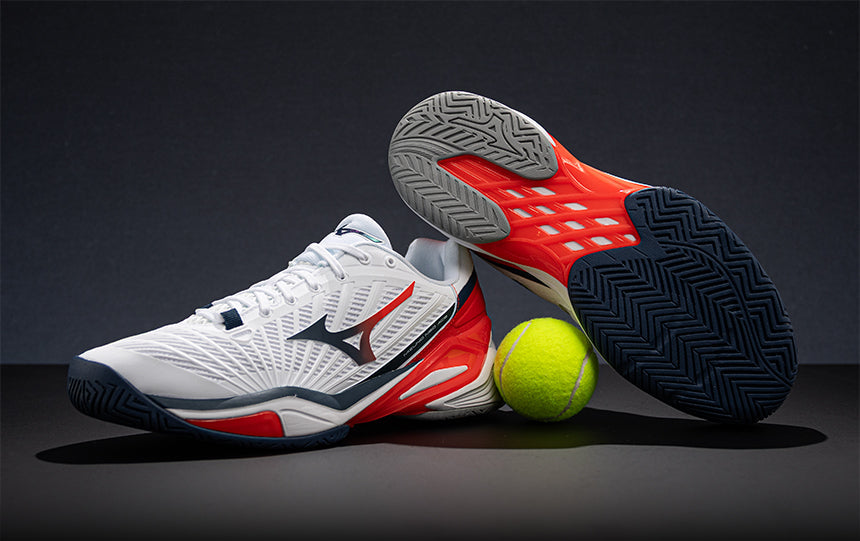 Tennis player wearing white, red and dark blue Mizuno Neo Ace tennis shoes on a blue court surface.