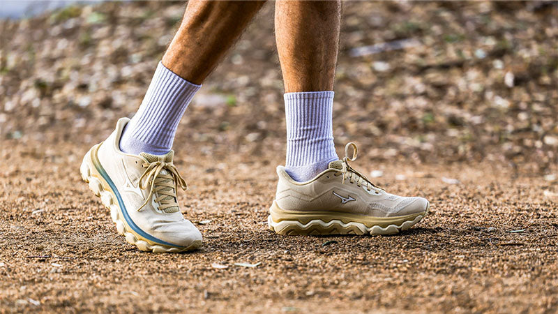 Person wearing khaki Mizuno Wave Horizon 9 running shoes with blue and gold accents and light grey crew socks on a dirt path.