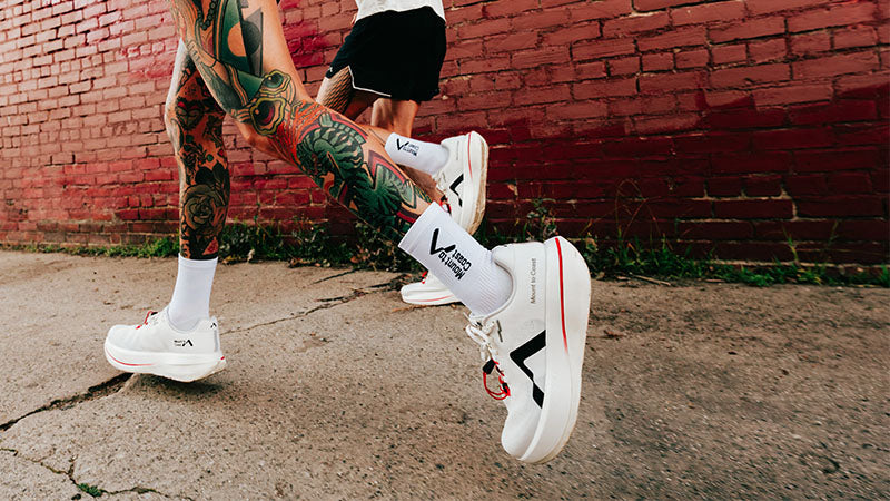 Two people running on a concrete sidewalk along a red brick building wearing white Mount to Coast C1 running shoes with black logos and red accents