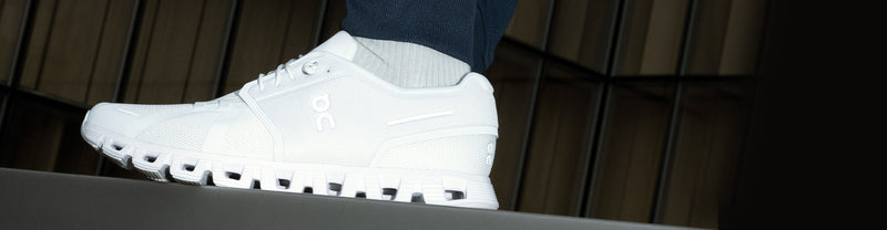Closeup of a person standing on a platform in On Cloud 5 All White running shoes against a dark background.
