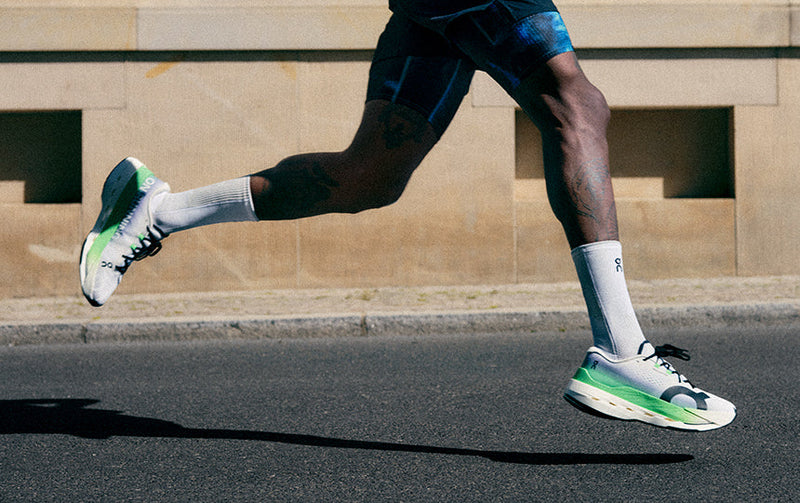 Person running on a street wearing white On Cloudboom Max shoes with green accents.