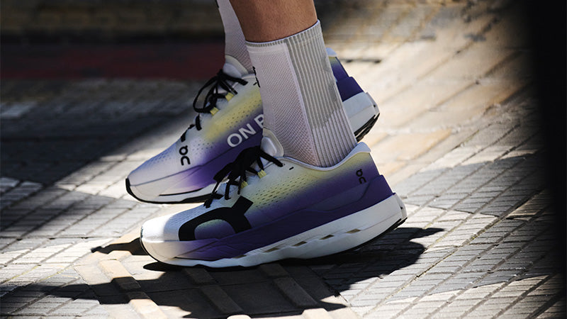 A person standing on a tile path in purple On Cloudboom Max shoes with white and yellow accents