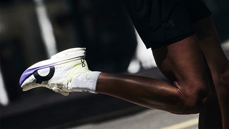 A person running in white On Cloudboom Strike shoes with a blurred city street in the background