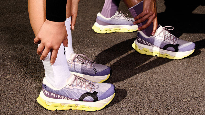 Two runners stretching on a paved surface in purple On Cloudmonster 3 Hyper running shoes with white and yellow accents and black "On" logos.