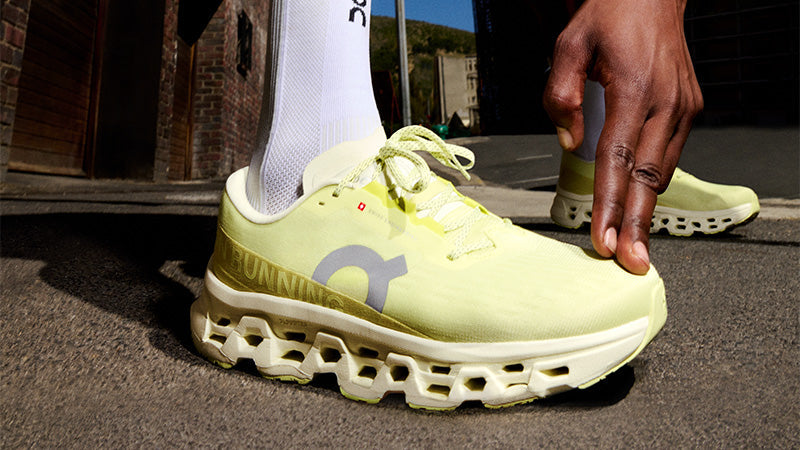 Closeup of a person wearing bright yellow On Cloudmonster 3 running shoes on urban pavement with brick buildings in the background.