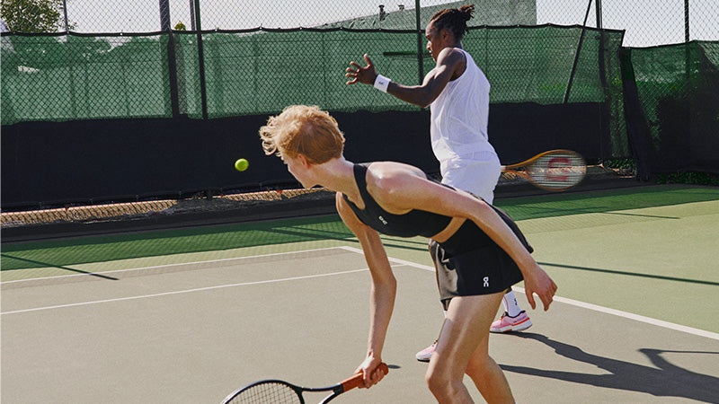 Two tennis players in action wearing On Court clothing with a green fence in the background.