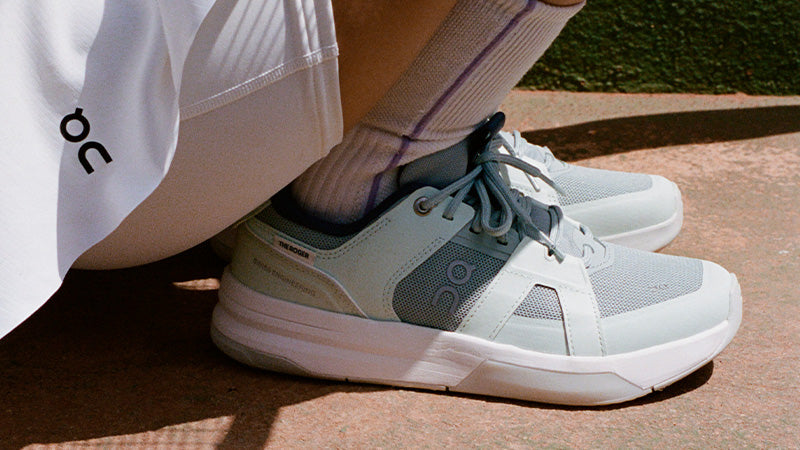 A close-up of a person wearing light blue and white On The Roger Clubhouse Pro tennis shoes with a white On skort and crew socks, sitting outdoors on a reddish-brown concrete surface.