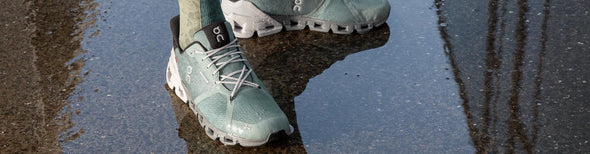 Man wearing On Cloudflyer Waterproof Sea/Glacier running shoes, standing in a shallow water puddle in an urban environment.