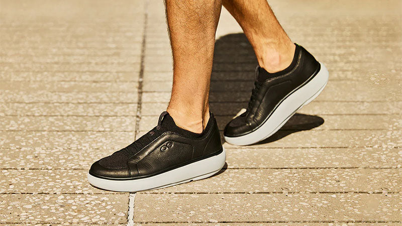 the feet of a person wearing black and white OOFOS Club+ Walking Shoes on a concrete brick surface