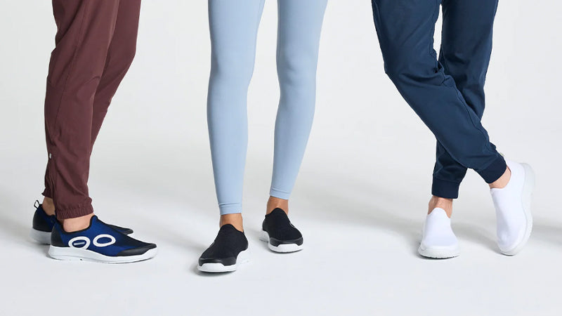 three people standing in differently colored OOFOS OOmg Sport recovery shoes on a white background