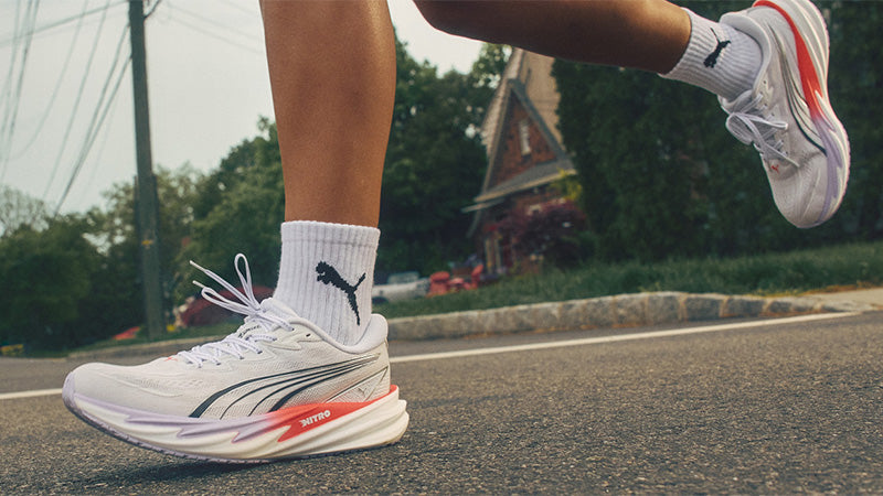 a runner on a paved road with a house and trees in the background, wearing white Puma Velocity NITRO 5 shoes with red and black accents.