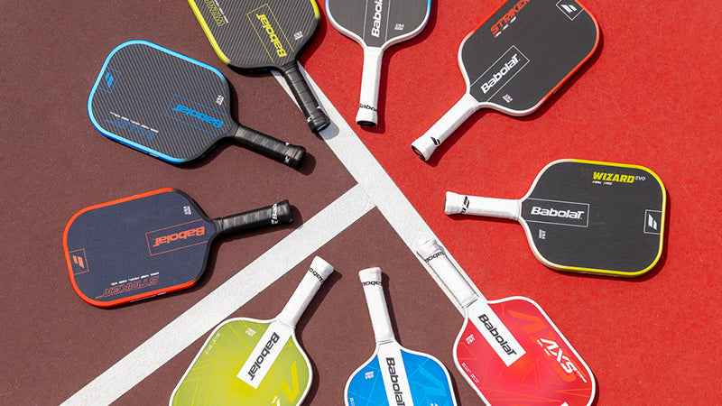 A collection of Babolat pickleball paddles in various colors and models arranged in a circular pattern on a red pickleball court background.