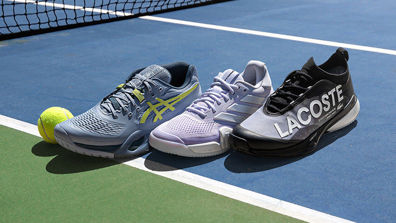 Three tennis shoes lined up on a blue tennis court next to a yellow tennis ball. The shoes are by ASICS, adidas and Lacoste, shown in gray/yellow, light purple, and black/white.