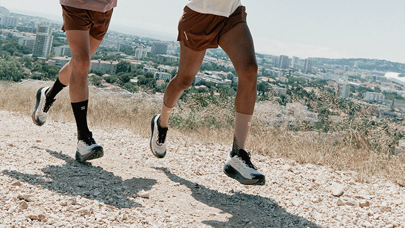 Two runners on a gravel mountain-side path above a cityscape wearing Salmon Aero Blaze 4 GRVL GTX running shoes