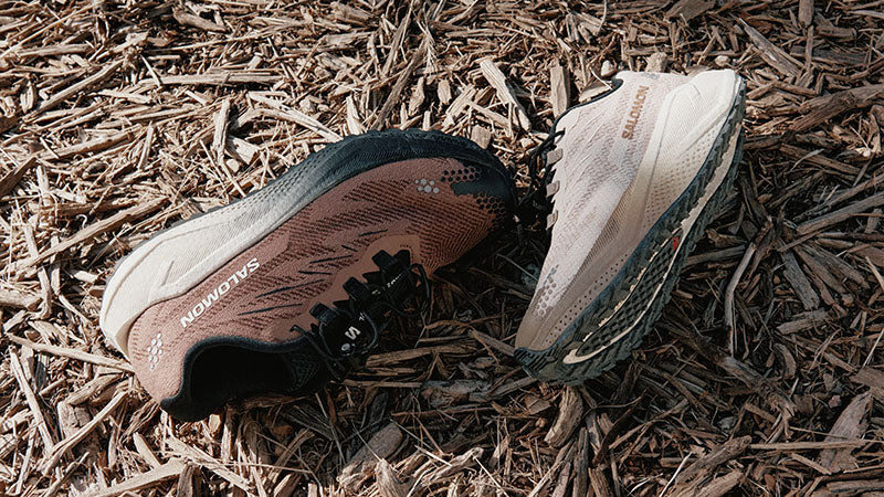 One men's and one women's Salomon Aero Blaze 4 GRVL running shoes shown on a bed of wood chips in an outdoor setting.