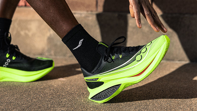 a person stretching on a sidewalk wearing bright green Saucony Endorphin Pro 5 running shoes with black and red accents.