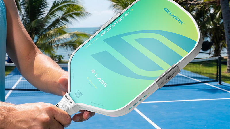 Closeup of a person on a blue, beachside pickleball court holding a Selkirk pickleball paddle with a blue-green and yellow gradient color treatment.