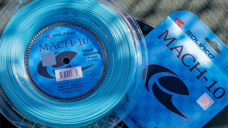 One reel and one set of Solingo MACH-10 tennis string in bright blue, placed on top of a tennis racquet on a tennis court surface.