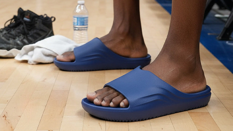 Closeup of a basketball player wearing blue Stand+ sandals while on the side of a court next to a towel, water bottle and a pair of black basketball shoes