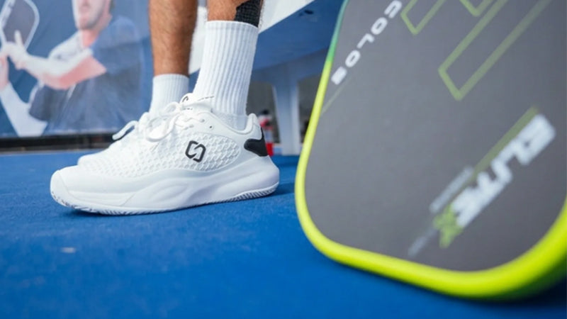 A close-up shot of a pickleball player wearing a white Stria Sport G1 Pro shoe with white socks, sitting on a bench along blue court next to a pickleball paddle.