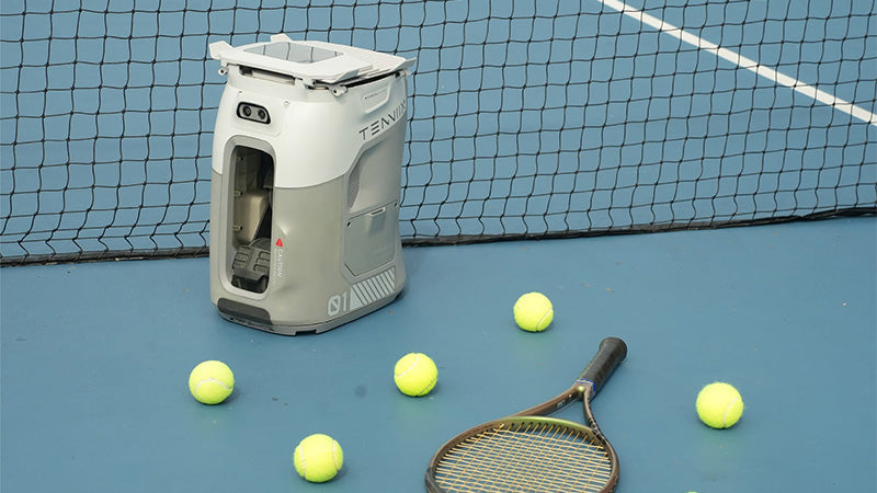 A white and grey Tenniix tennis/padel ball machine sitting on a blue tennis court with a net behind it, several yellow tennis balls around it, and a tennis racquet in the foreground.