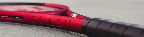 Image of a red Wilson Clash v2 tennis racquet, focused on the neck, laying flat on an outdoor tennis court.