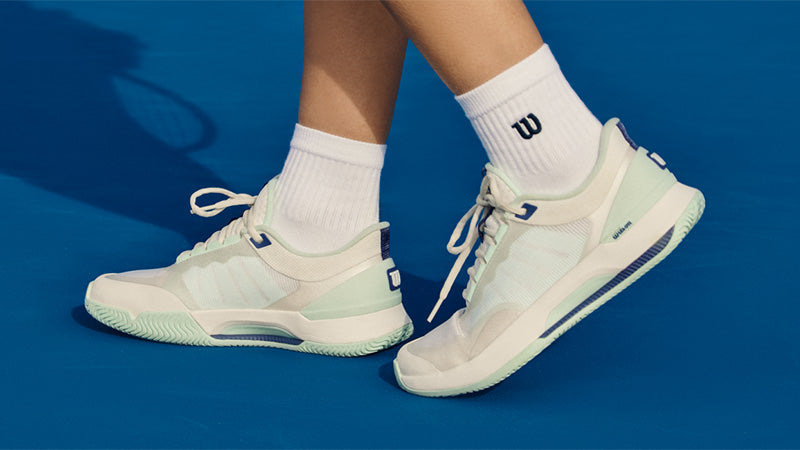 A tennis player standing on a blue court surface wearing white and light green Wilson Intrigue Lite women's tennis shoes with dark blue accents