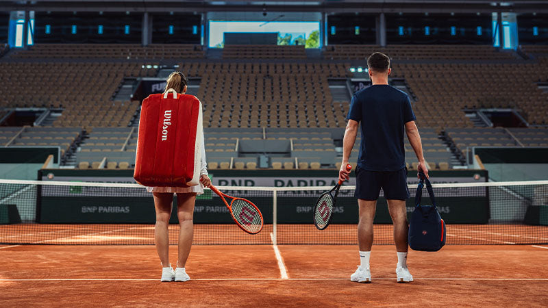 Two tennis players standing on a clay court inside an empty stadium with Wilson Roland Garros 2026 racquets and bags.