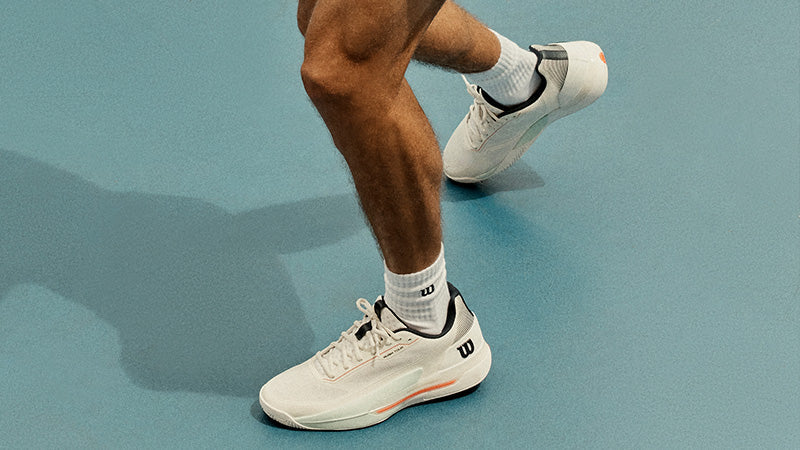 Person wearing white Wilson Rush Tour 5 tennis shoes on a blue court
