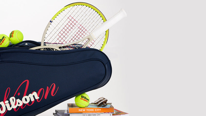 Wilson US Open racquets sticking out of a matching bag with balls and books about New York City against a light gray background.