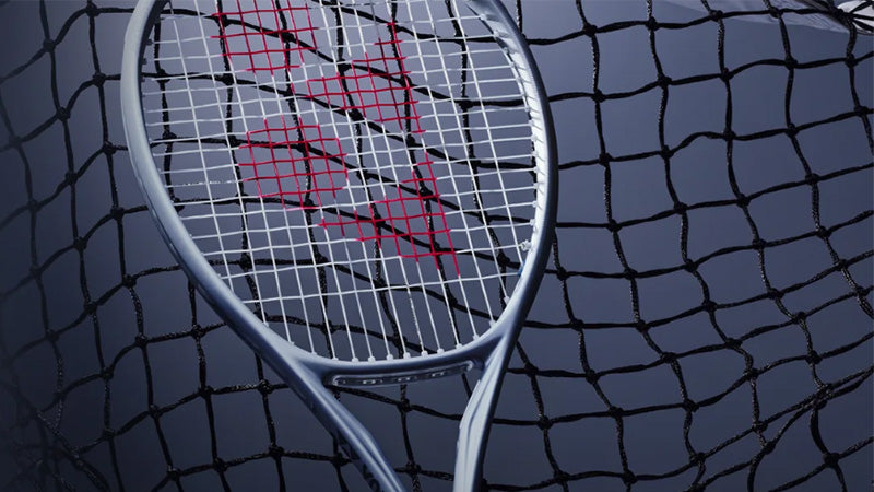 A silver Yonex Muse tennis racquert leaned against a net on a blue-gray tennis court.